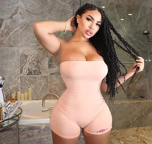 Sexy woman with black hair stands in a pink strapless bodysuit to show off bust.
