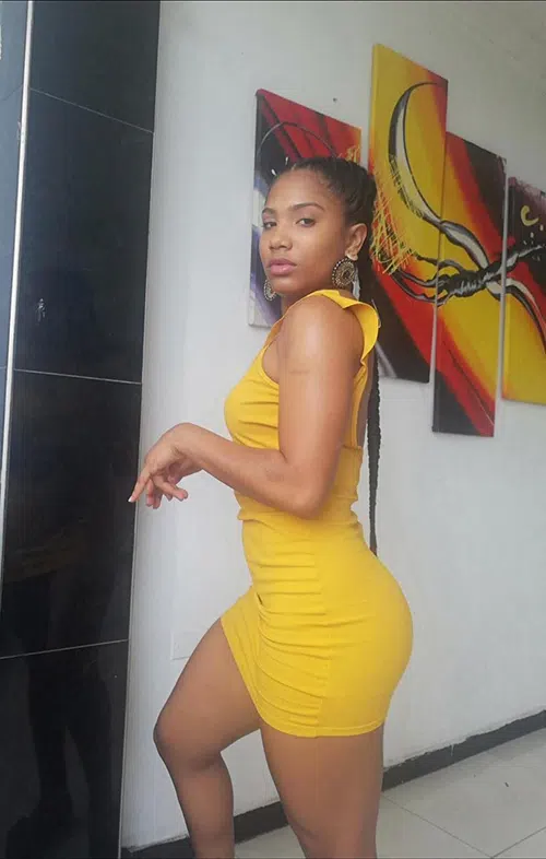 Black girl with long braid poses in a skintight yellow dress in an art gallery.